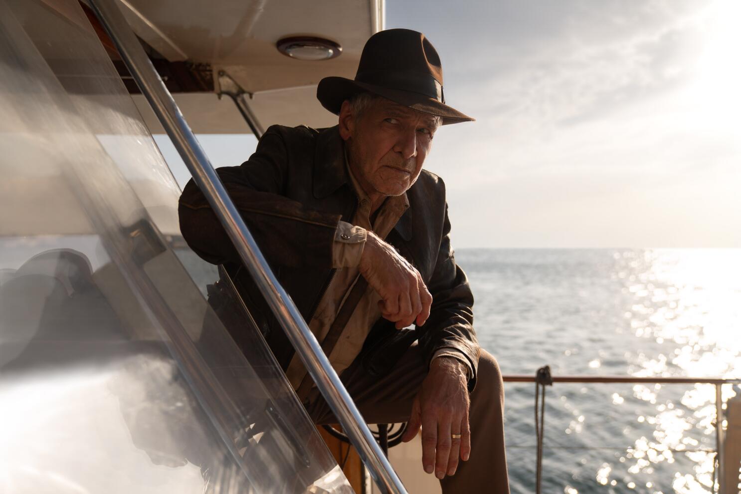 Indiana Jones con su chaqueta de cuero café y una fedora a juego. Posa con una pierna apoyada en un barco.