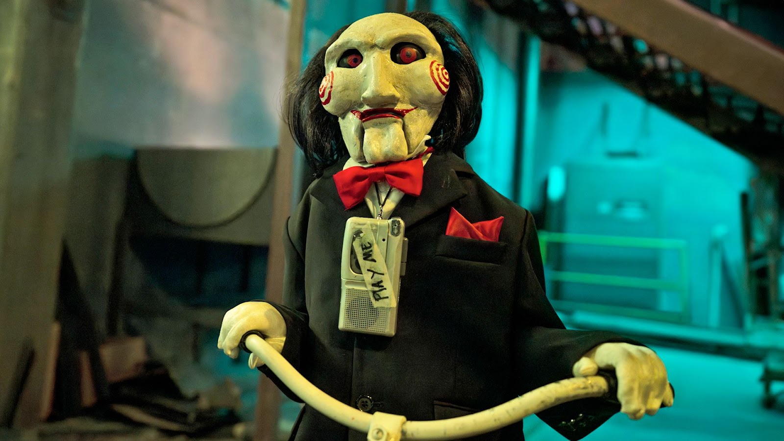 El personaje Billy de la película de terror Saw, está andando en triciclo con su clásico esmoquín negro, humita roja y una máscara blanca con detalles de espirales en las mejillas.