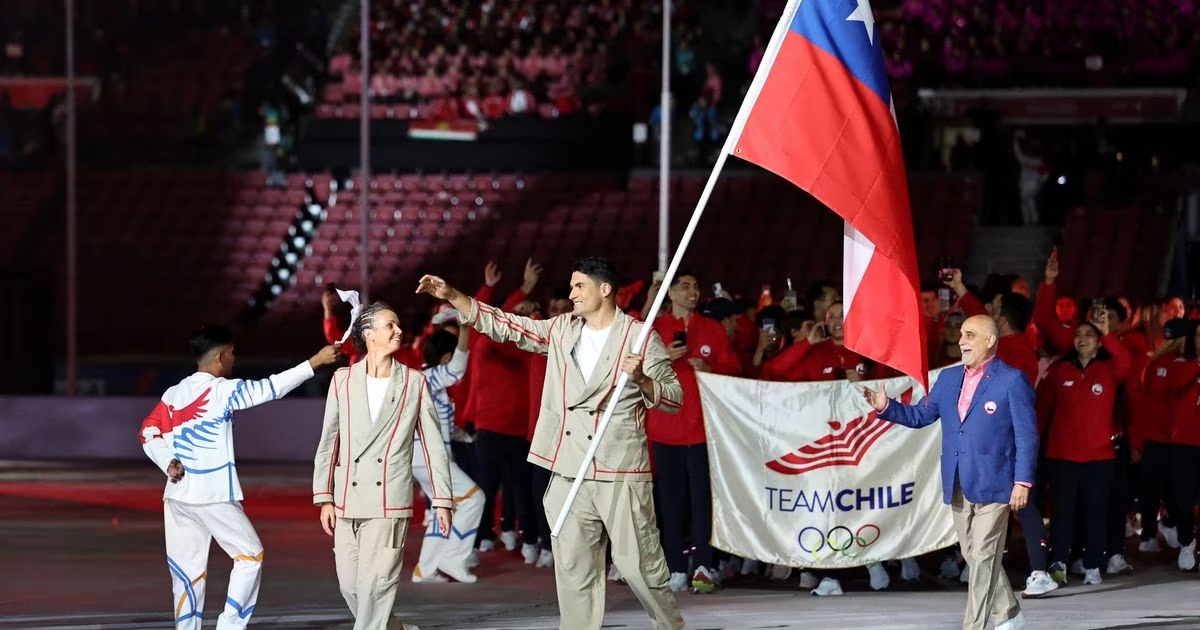 Los abanderados del Team Chile, Kristel Köbrich y Esteban Grimalt, llevando la bandera y encabezando el desfile de la selección de deportistas chilenos en la inauguración de los Panamericanos 2023. Llevan un traje caqui con líneas rojas.