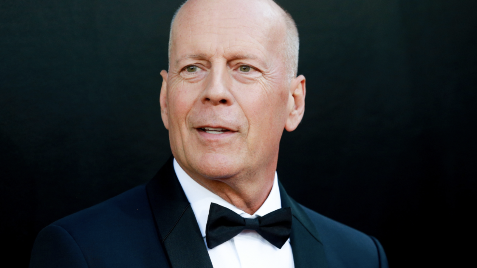 Bruce Willis en un fondo negro. Viste un traje de color azul marino con una camisa blanca abotonada, al centro tiene puesto un corbatín.