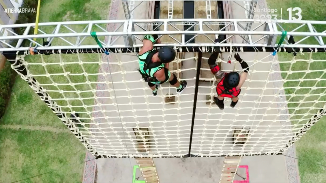 Dos participantes, uno del team verde y otro del team rojo, usan casco negro y se ven descendiendo de un muro de escalada con redes.