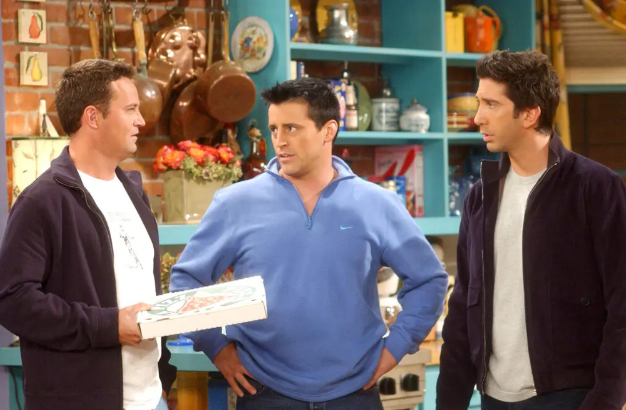 Joey y Ross miran a Chandler Bing mientras él sostiene una caja de pizza