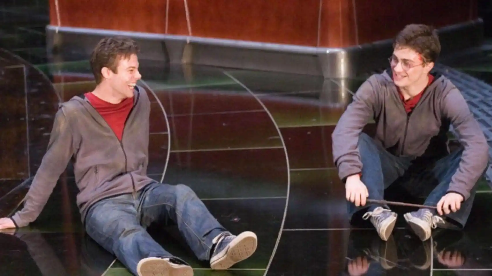 Daniel Radcliffe junto a David Holmes. Ambos llevan puesta la misma ropa interpretando a Harry Potter. Lucen un par de jeans, zapatillas azules, polerón con cierre color café y una polera roja. Están sentados en el suelo.