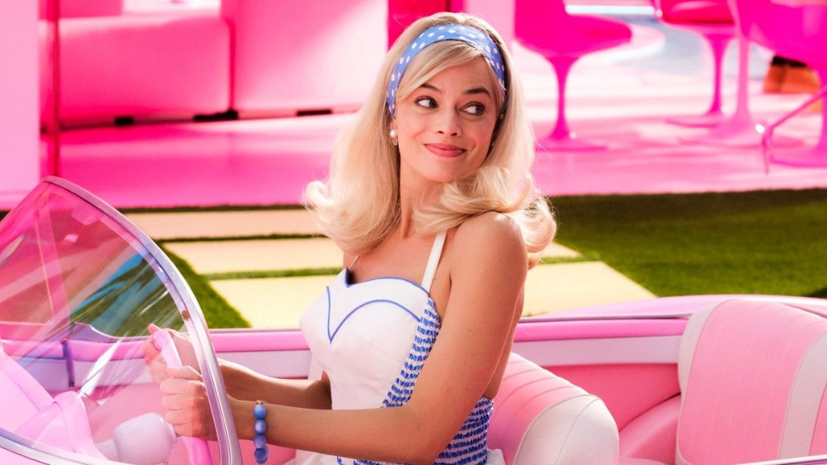 Margot Robbie interpretando su papel como Barbie. Lleva el cabello rubio suelto y con un cintillo azul con lunares blancos, el que hace juego con su vestido de tirantes. Se ve a Barbie manejando su auto descapotable rosado.