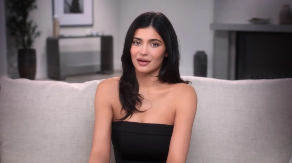 Kylie Jenner sentada en un sofá, vistiendo un top sin tirantes negro, hablando en una entrevista en un entorno interior moderno y elegante.