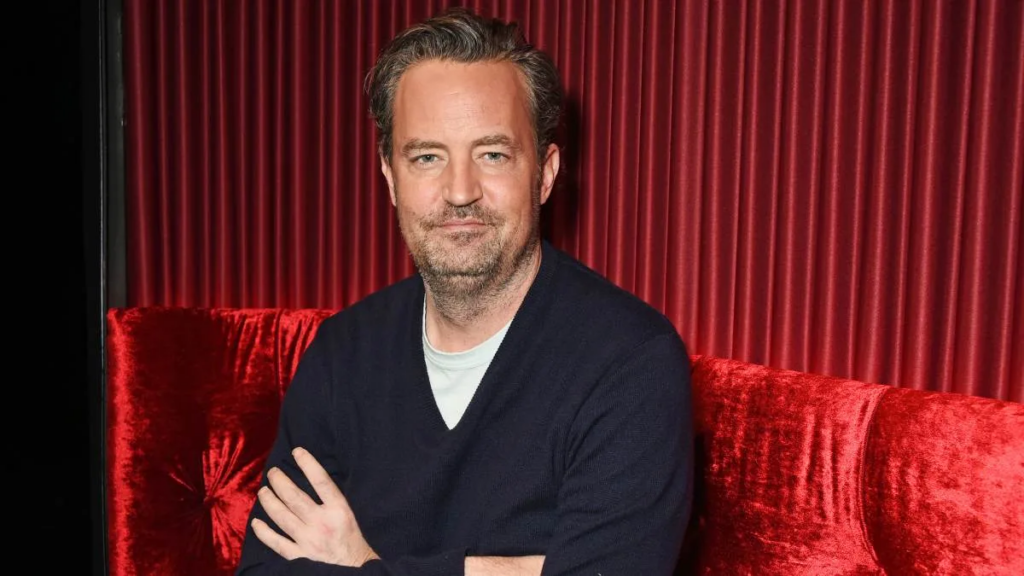 Matthew Perry sentado en un sillón rojo, con los brazos cruzados y sonriendo levemente durante una entrevista, luciendo relajado y casual.