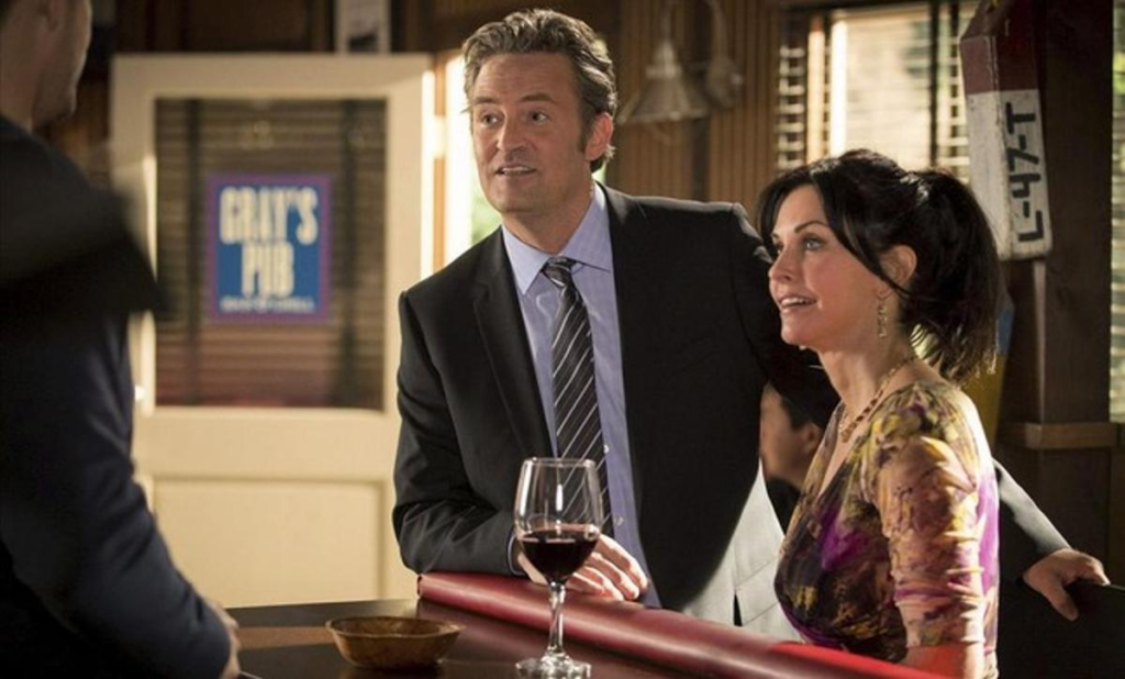 Matthew Perry y Courteney Cox en una escena juntos, ambos vestidos elegantemente, con Matthew sosteniendo una copa de vino en un bar.