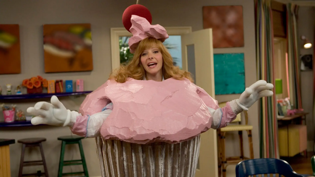 Lisa Kudrow vestida de cupcake en una escena humorística, con un traje rosa y esponjoso, haciendo un gesto exagerado y divertido.