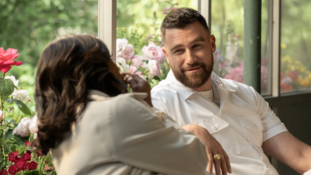 Travis Kelce con barba, vestido con camisa blanca, sonriendo mientras conversa con una mujer en un entorno al aire libre rodeado de flores coloridas.